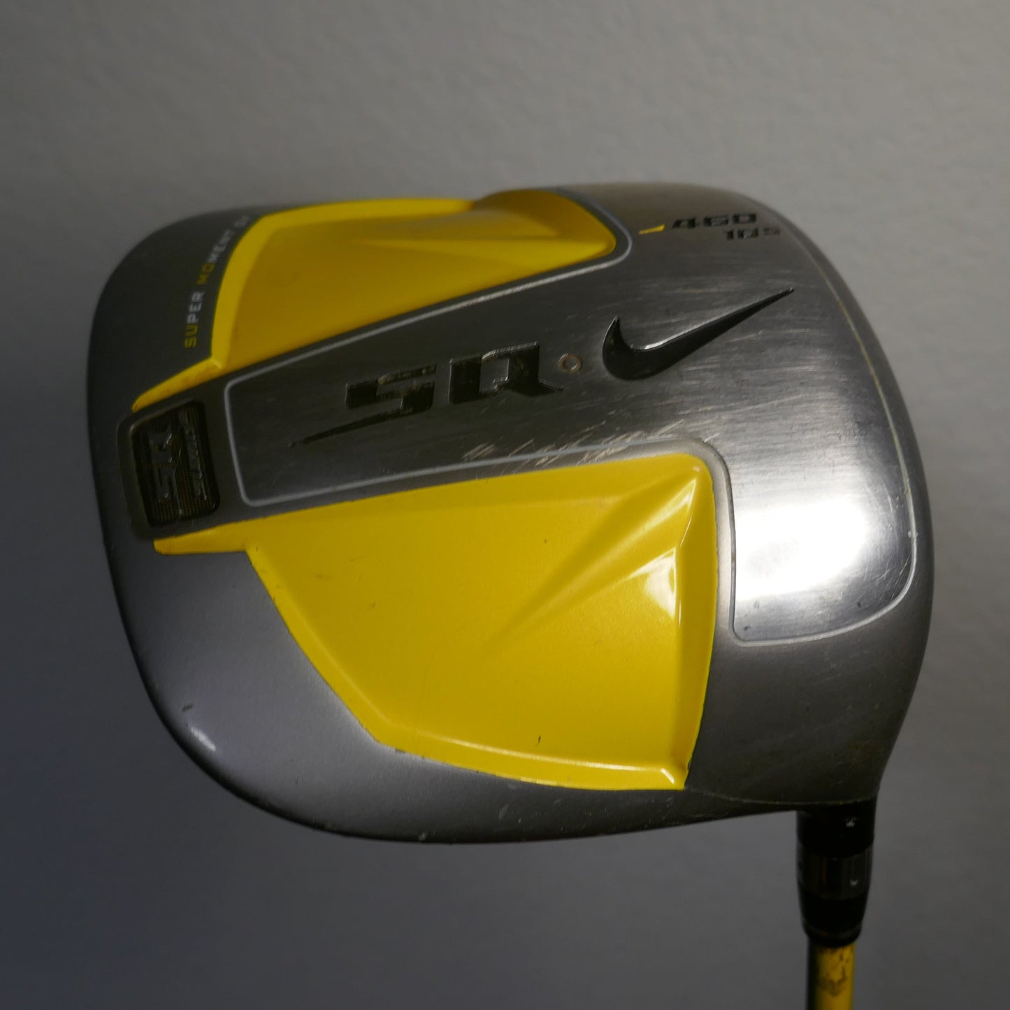 A Nike golf driver with a yellow and silver head, featuring the Nike swoosh and 'SQ' branding, shown against a plain gray wall.