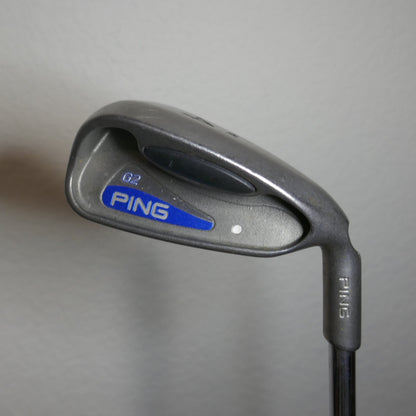 A Ping golf iron club head and part of the shaft, photographed against a plain light gray wall. The club face shows the PING logo in blue and white, and the model is G2.
